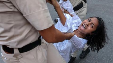 Two uniformed men drag a woman she appears to be shouting. She is wearing a white shirt and has long, black hair.