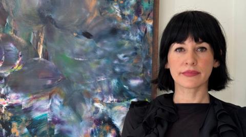 A woman with black hair in a bob and red lipstick. She is looking at the camera and standing in front of a painting which is in blues and greens, marked in differing directions.