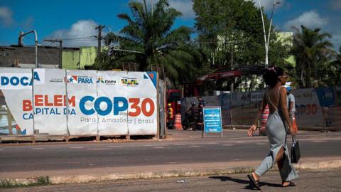 A woman walks past an infrastructure project under way for COP30 in Belem. She has her hair up and is wearing a grey dress and carrying a black bag. Misaligned posters in white, blue and orange read "Belem Capital Da COP30".