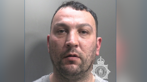 A mugshot of Edwards with a stubbly, black beard with short black hair. There is a North Wales Police logo in the bottom right-hand corner.