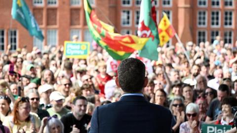 The back of a man's head is shown, with hundreds of people in front of him, many waving Welsh flags