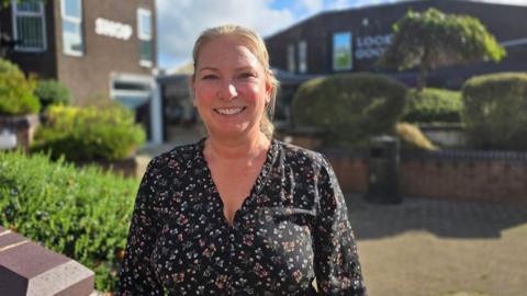 Sally Hellwich has blonde hair tied back and is smiling. She's wearing a black blouse with a colourful floral pattern. She is standing outside in a pedestrianised street with bushes and trees around brown buildings, on a sunny and cloudy day.