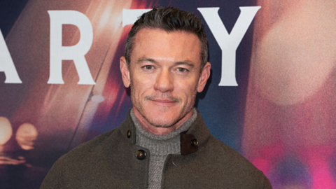 Man with short grey/brown hair and facial hair, wearing grey jumper and brown jacket and looking dorectly at camera.