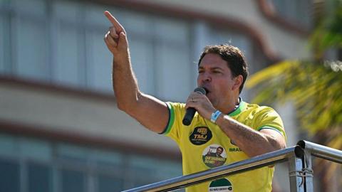 Alexandre Ramagem points with his finger with one hand while holding a microphone in the other hand as he is speaking at a demonstration in support of former Brazilian President Jair Bolsonaro at Copacabana Beach in Rio de Janeiro on 3 August, 2025. He is wearing a Brazil football shirt with several stickers on it. One bears a photo of ex-President Bolsonaro.
