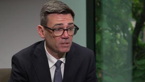 Andy Burnham, 56, who has greying brown hair and is wearing glasses, speaks to the camera. He is sitting in a brown chair, by a window, and is wearing a dark suit.