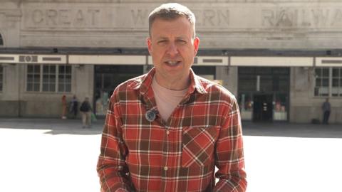 Daniel Davies, who has blonde hair, in a chequered red and dark red shirt stood outside a station