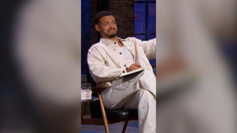 A man with short hair, a moustache and beard is sitting in a chair. He is wearing a white vest, jacket and trousers.
