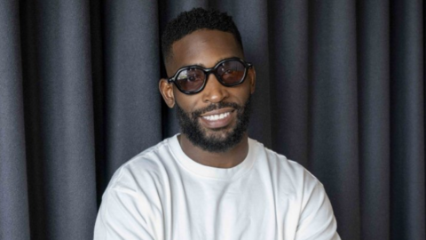 A man smiling at the camera. He is wearing sunglasses and a white top. His arms are folded, and he is standing in front of a grey curtain.