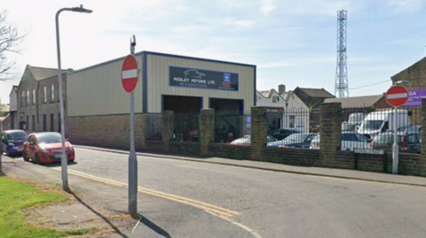 A street corner with cars parked on the street on the left. To the right is a motor company with cars parked behind a gated wall.