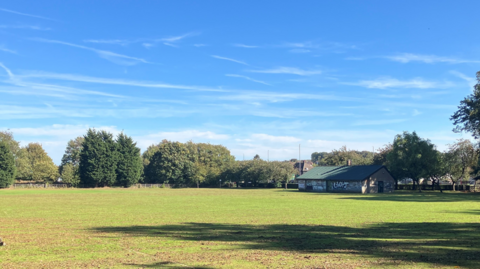A large area of green space with a building and trees in the background and blue skies.