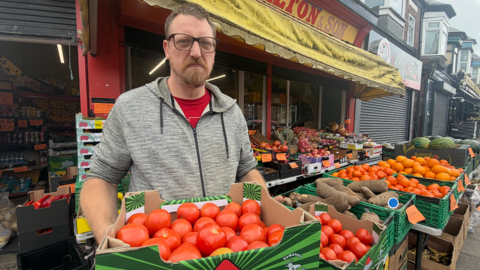 Brad Purnell, who is wearing large black glasses a red shirt and a grey fleece, is holding a green cardboard box with more than dozen tomatoes. In the background are several green boxes with potatoes, oranges and melons. He is in front of his shop which features a red front with a yellow awning, and a sign which reads J Charlton & Son.