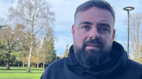 A man with black hair and beard who is wearing a black hooded top. There is a park behind him with trees and grass and someone is walking a dog.