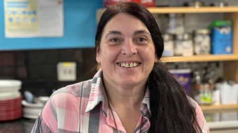 Patsy Lockerby, who has long dark hair in the ponytail and is wearing a pink, white and grey checked shirt. She'd standing behind the counter in her tearoom.