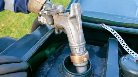 A gloved hand holds a home heating oil pump over a domestic oil tank