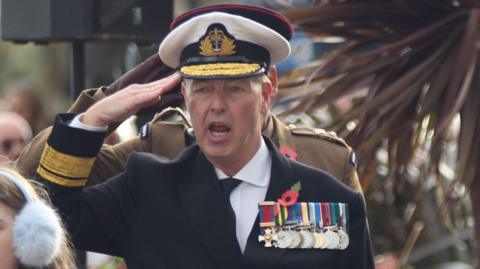 A man dressed as an admiral gives a salute. He wears a white hat, navy jacket, white shirt and tie. On his chest are a series of medals hung by colourful ribbons.