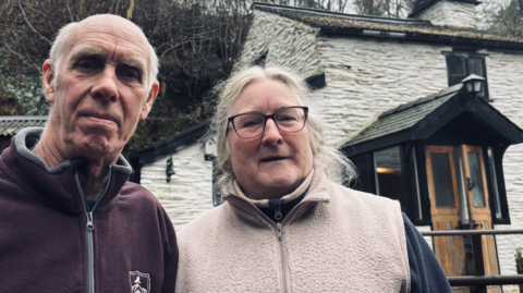 Jill and Ross Darbyshire, an older couple wearing fleeces, stand outside their cottage home. Ross is bald with short white hair on the sides, while Jill's white hair is tied back, she is wearing glasses. The cottage is whitewashed with a black wooden porch.