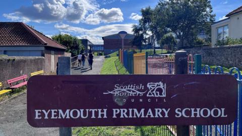 A maroon sign saying Eyemouth Primary School in white lettering with colourful railings and buildings in the background
