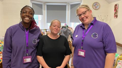 Three women stand in a room with baby clothes on a rail behind them and toys and baby toiletries on tables. Two women are wearing purple and the woman in the middle is wearing a black T-shirt.