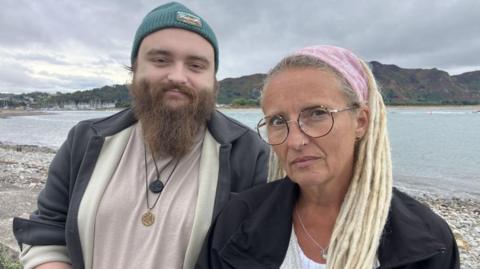 Ryan Shields and his mother Aly Shields standing together looking towards the camera. His mother is wearing a pink bandana on her blonde hair and has brownish glasses. Ryan is wearing greyish jacket and a blue beanie. He also has a long, light brown beard.