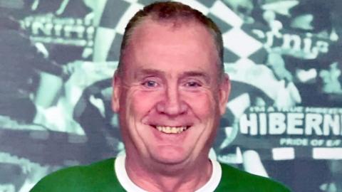 A man is smiling at the camera. He is standing in front of a poster that says Hibernian football club. He is wearing a green and white t-shirt. He has short brown receding hair.