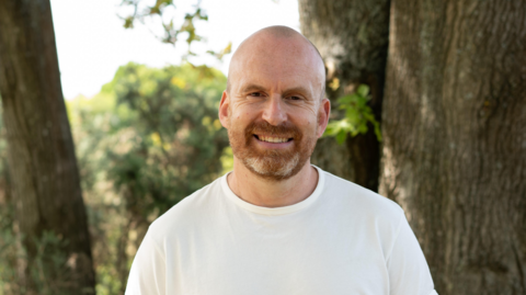 Matt Haig is wearing a white T-shire and is stood outside. He has a beard and is smiling at the camera.
