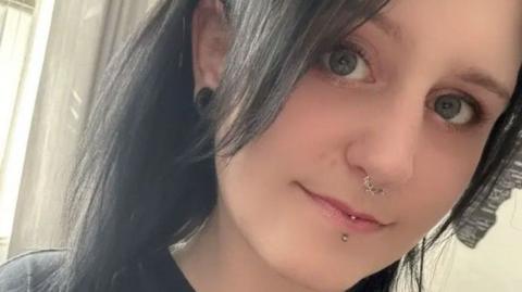 A woman with dark hair pictured from the shoulders up. She has a nose and lip piercing and is wearing a black T-shirt.