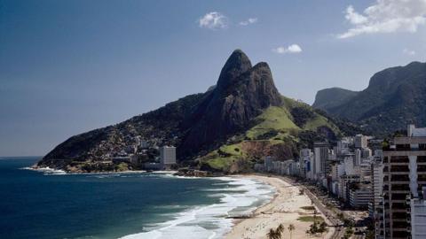 Wide shot of Ipanema Beach in Rio De Janeiro with Morro Dois Irmaos at the end. To the left of the frame, is a row of residential buildings, to the left is the sea with waves crashing into the beach