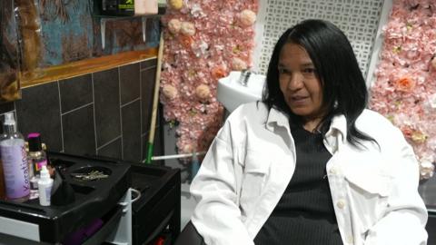 A woman sits in a salon with pink flowers behind her. Hair products can be seen on a stand to the left of her.