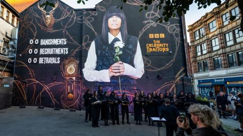 A mural featuring a stag and Claudia Winkleman. She holds a white flower in a tunic dress. There is an adjacent wall with portraits on it and a choir stands in front of the building.