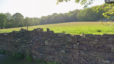 A field with a slightly broken dry stone wall in front of it and trees around the edge.