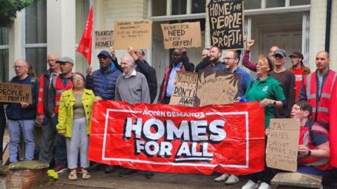 A group of people standing outside two converted houses holding a banner that reads 'Homes for All'. Others are carrying cardboard placards.