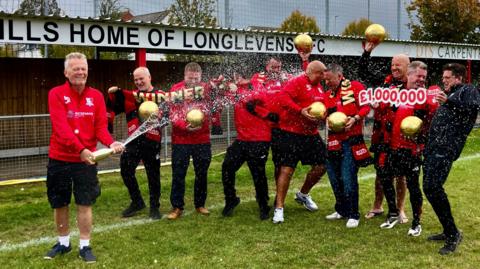 Ten members of a football club - most of whom are middle aged men - celebrate their National Lottery win on their pitch. They are all wearing a red club fleece and holding gold coloured footballs as one sprays champagne from a golden bottle over the others. Some of the members are holding red and black scarves which say "winner" in gold lettering and small signs that read "£1,000,000".