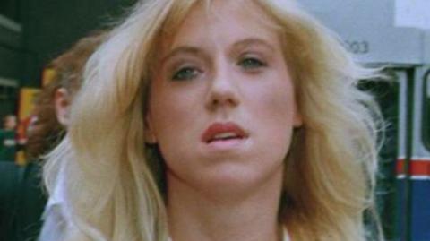 Tracie Andrews with long blonde hair. She is wearing a white shirt with a black jacket.