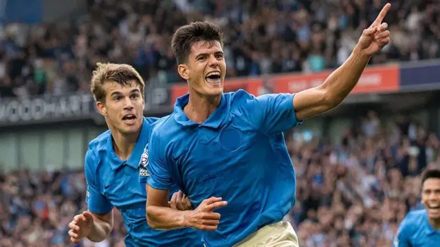 Chema Andres, wearing a blue shirt, celebrates scoring with his left index finger outstretched, and teammate Luka Vuskovic running to his right