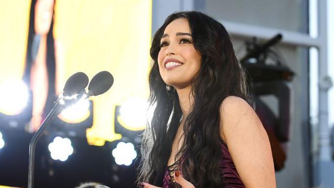 Valkyrae at a gala event in LA in 2024. She has long dark hair worn loose and wears a burgundy dress. She smiles and looks up as she delivers a speech on stage.