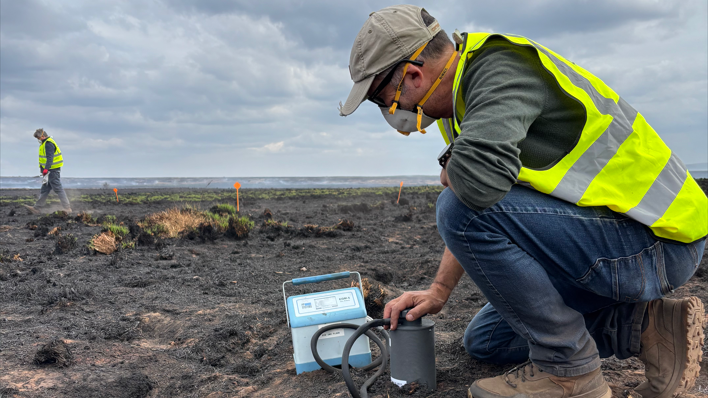 Professor Dave Chadwick from Bangor University sampling damaged remains following wildfires at Mynydd Llangynidr in Bannau Brycheiniog. He is wearing a cap, high vis jacket, mask and boots.