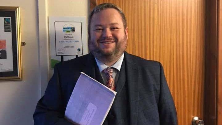 Gareth Ritter pictured smiling and wearing a dark suit and a matching waistcoat and patterned tie holding a purple exercise book. He has short brown hair and facial hair.
