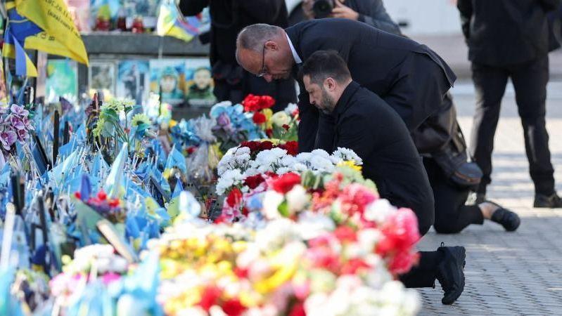 Merz and Zelensky knee on the ground as they lay flowers. In front of them is a display of flowers in shades of reds, whites, yellows and blue. Both men are wearing dark clothes.