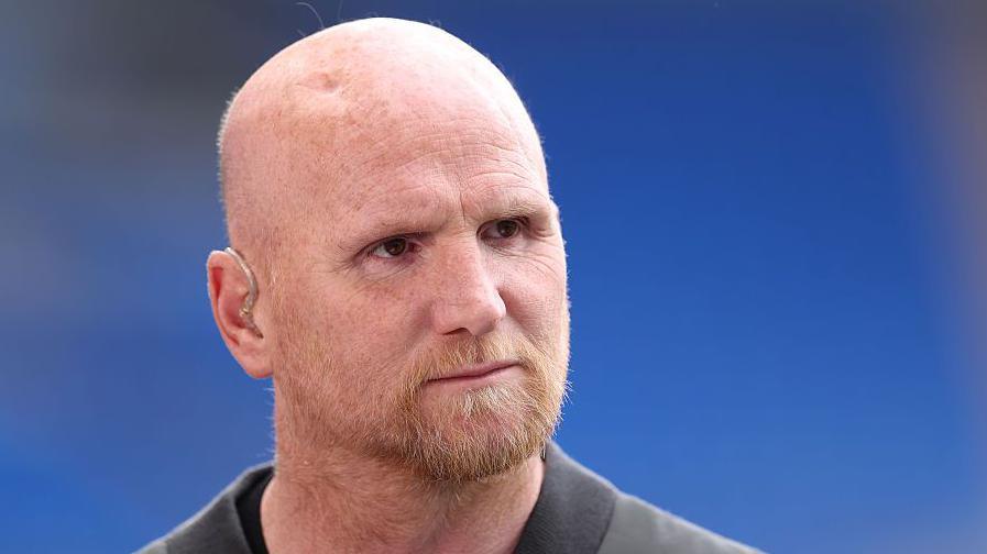 John Hartson has a bald head and a ginger beard. He has an earpiece in his ear. He is looking off camera and there is a blue background