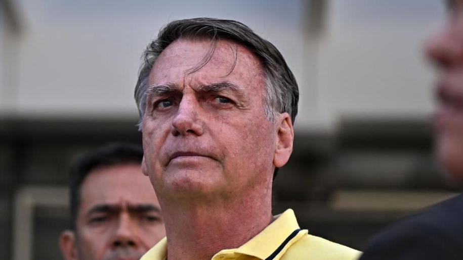 Jair Bolsonaro, wearing a pale yellow polo shirt, looks ahead as he leaves a branch of the DF Star hospital services in Brasilia, Brazil, on 16 August 2025. His brow is furrowed and two strand of hair are falling onto his forehead.
