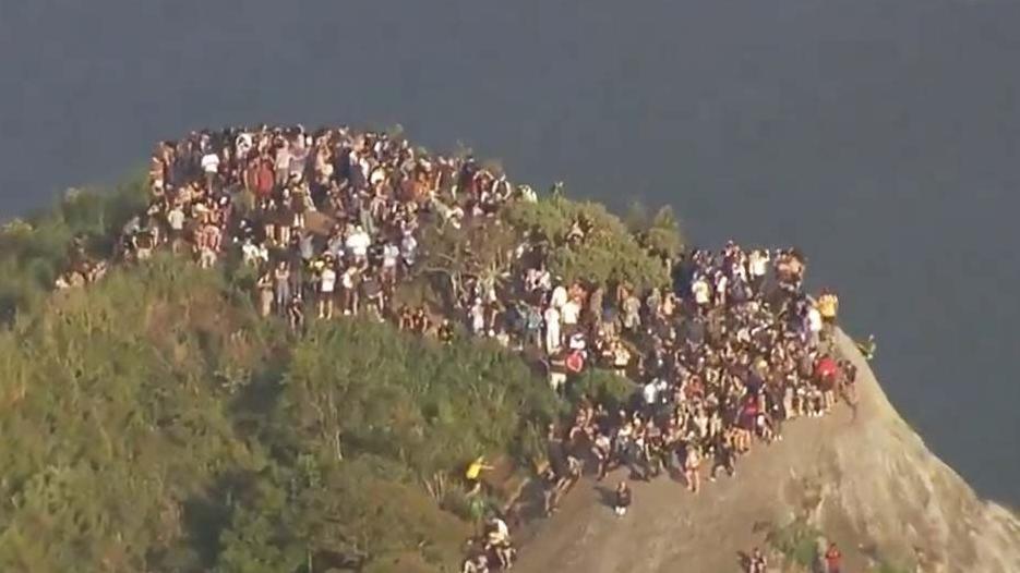 Aerial photograph shows a large group of people standing at the top of a high point in Rio de Janeiro, Brazil. It's early morning, and the shadows are long, indicating sunrise.