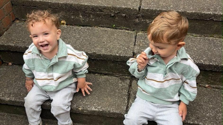 Noah (left) and his brother Malik (right) both sit on concrete steps in matching outfits. Noah has light brown riglets and looks up at the camera smiling. Malik looks down at the step.
