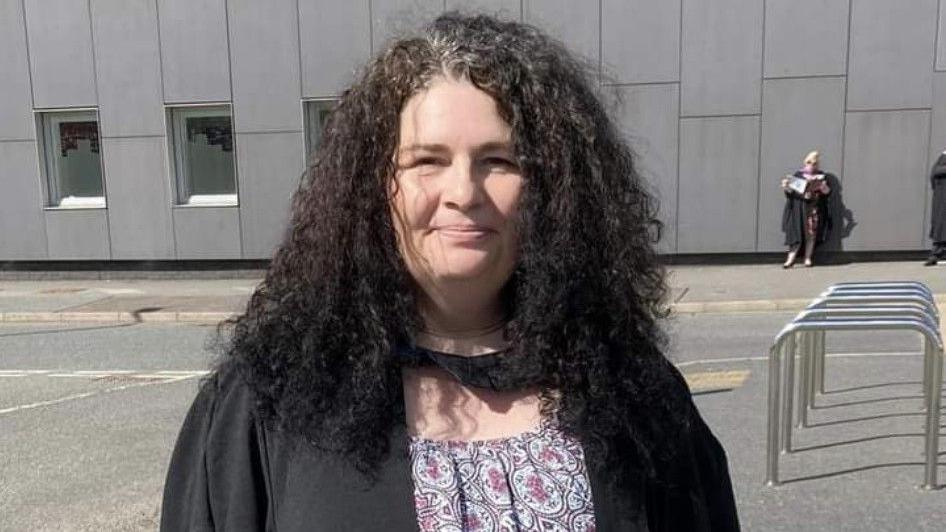 Diane Barnes is a middle-aged woman standing outside a large grey building. She is wearing a graduation gown and has black hair. She is smiling at the camera.