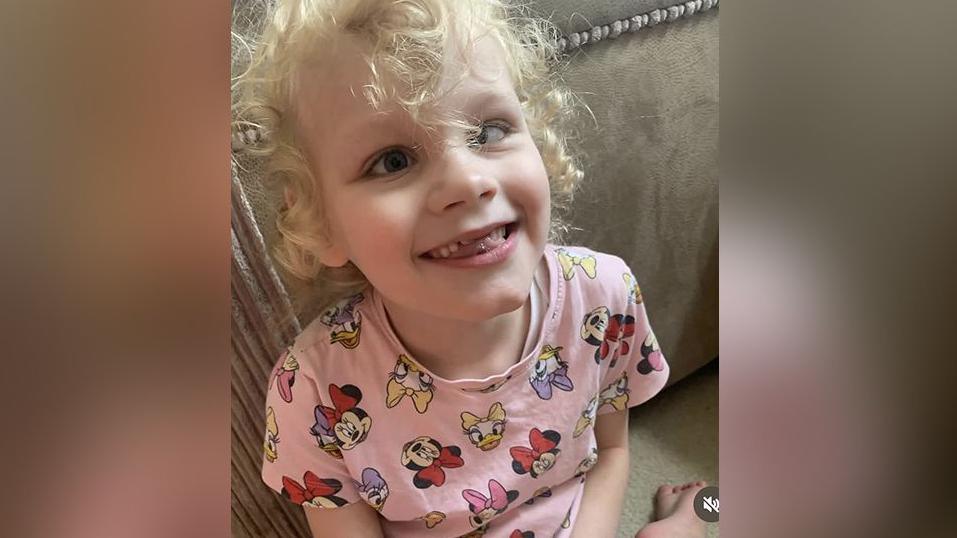 Seven-year-old Isla with blond curly hair wearing a pink Disney patterned t-shirt sits on the floor leaning on a beige couch. She is smiling.