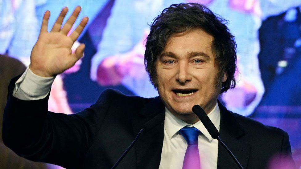 Javier Milei wearing a suit and surrounded by people, waves to a crowd with his mouth open