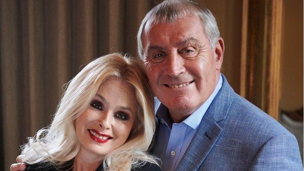 Peter Shilton, who is wearing a blue suit, has his arm around Steph Shilton, who has shoulder-length blonde hair and is wearing a black top.