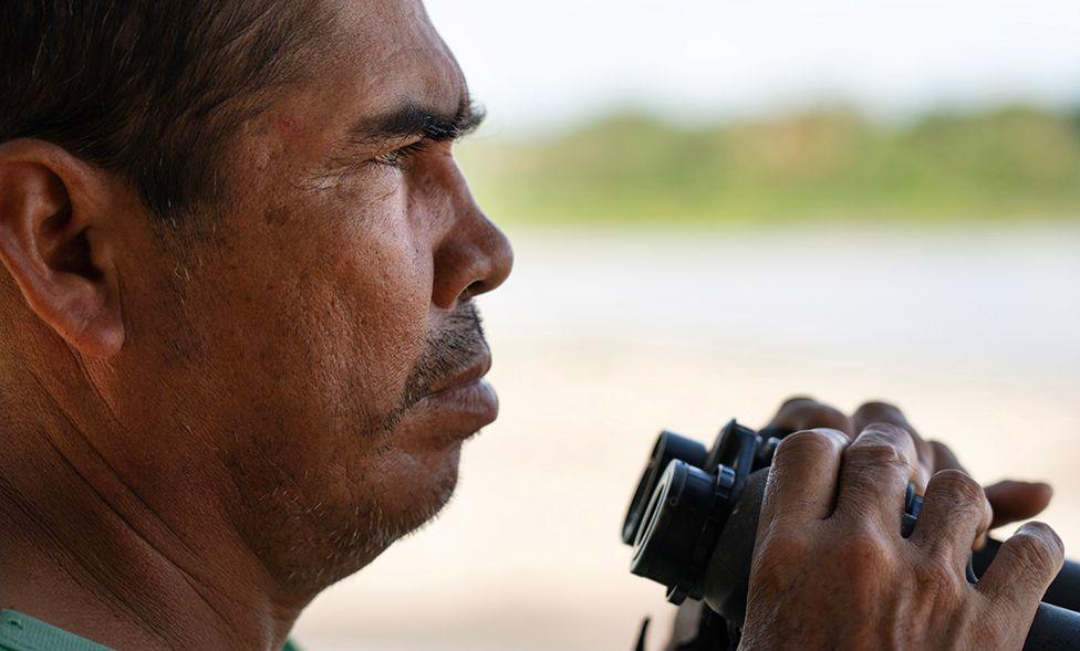 Close-up of Antonio holding black binoculars with both hands, positioned near a body of water. The background is out of focus, showing light-colored sand and greenery in the distance.