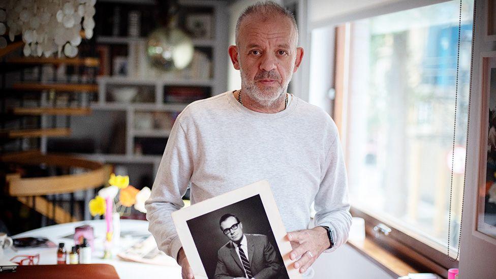 Antony Easton wearing a light-colored long-sleeve shirt stands indoors, holding a black-and-white photograph of his father, Peter Easton, dressed in a suit and tie. Behind him is a room with bookshelves and various items, lit by natural light from a window on the right.