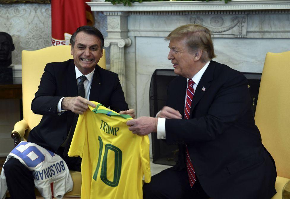 Bolsonaro gives Trump a yellow Brazil soccer jersey with the name TRUMP on the back. Trump is smiling as he receives it, seated in the Oval Office.