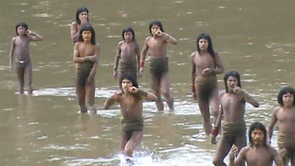 A group of Mashco Piro tribesman wading shin-deep in a river, calling out to someone on the other side. They are wearing only loin cloths and appear to be trying to catch someone's attention.